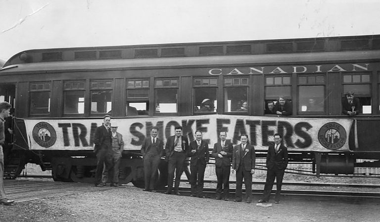 Black and white photograph of train with “Trail Smoke Eaters” banner and team posing in front of train