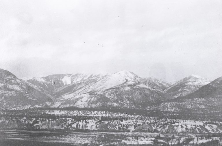 Black and white photograph of valley and mountain in background covered in snow. 